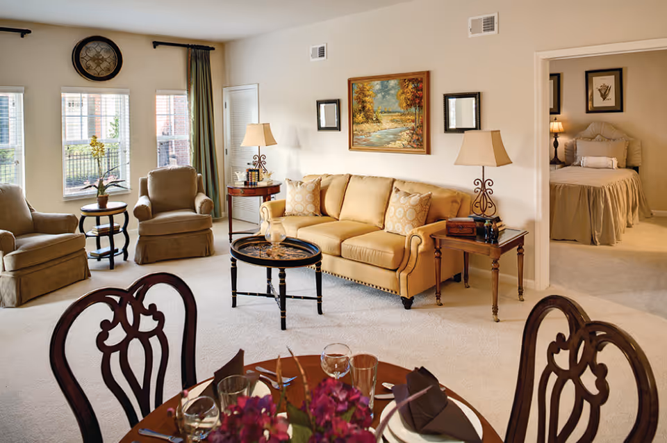 A cozy living room with a yellow sofa, two armchairs, a round coffee table, and two side tables with lamps. There is a painting on the wall above the sofa and a clock above the windows. The room has large windows with green curtains. In the foreground, a dining table is set with plates, glasses, and a floral centerpiece. A bedroom with a bed and framed pictures is visible through an open doorway.