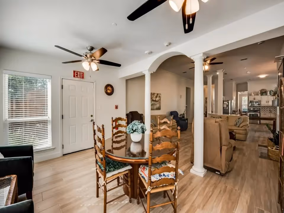 Interior view of a senior living facility showing a dining area with a round glass table and wooden chairs, a living room with beige armchairs and a blue recliner, ceiling fans with lights, white columns and arches, and a kitchen in the background with stainless steel appliances and white cabinetry.