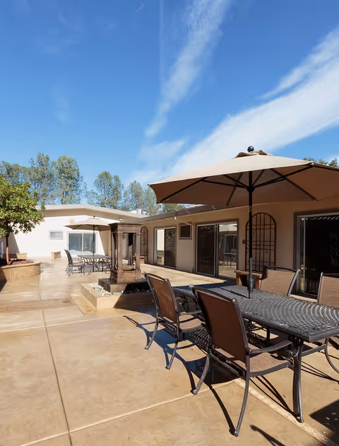 Outdoor patio area at Rock Creek Care Center with metal tables and chairs, large beige umbrellas providing shade, a decorative water fountain, and a clear blue sky with some clouds. The patio is surrounded by a single-story building with large windows and doors.