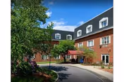 Front entrance of a red-brick senior living building with a covered drop-off, curved driveway, and landscaping under a blue sky.