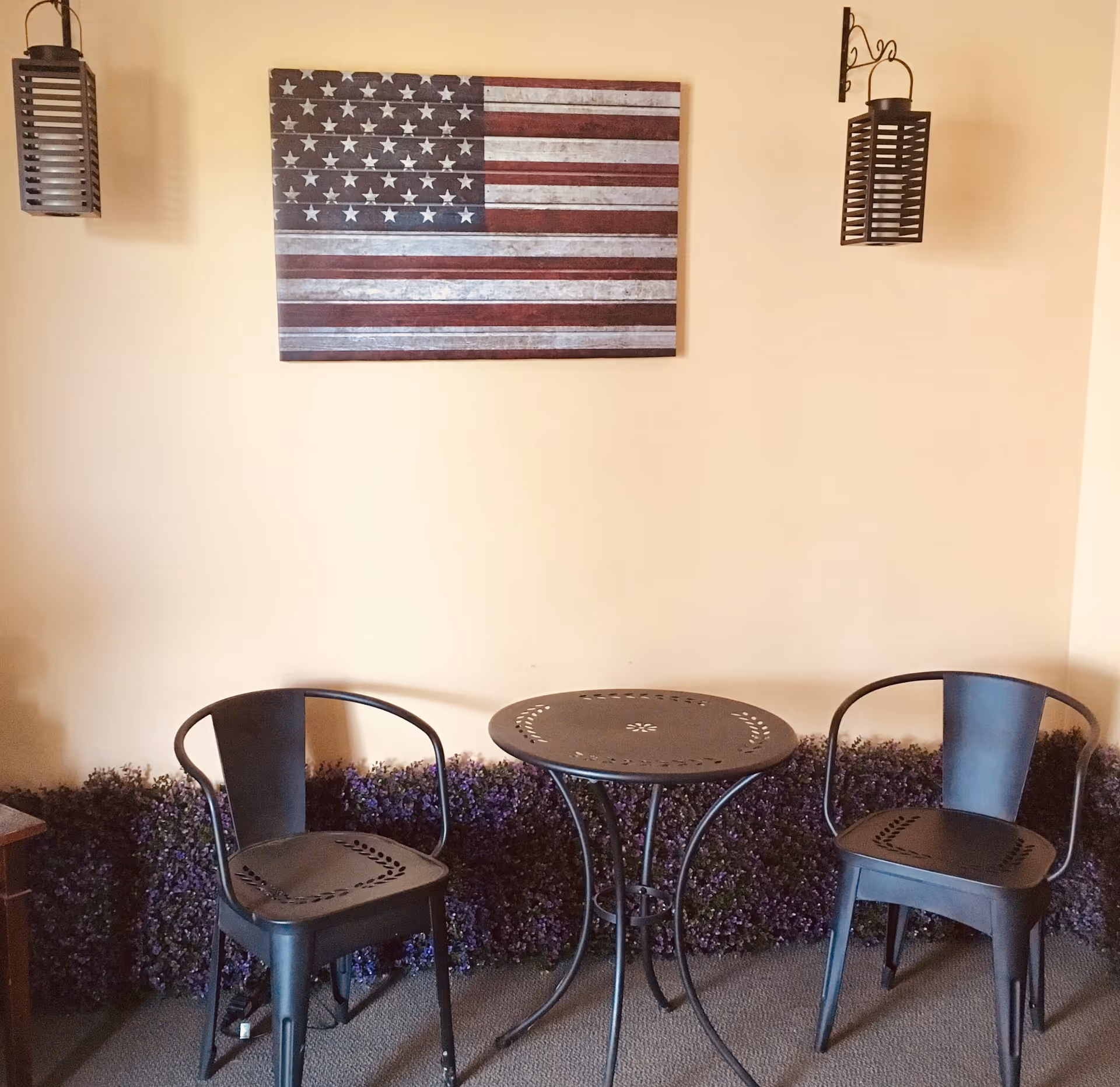 Small indoor seating area with two metal chairs and a round metal table beneath an American flag wall art and hanging lanterns.