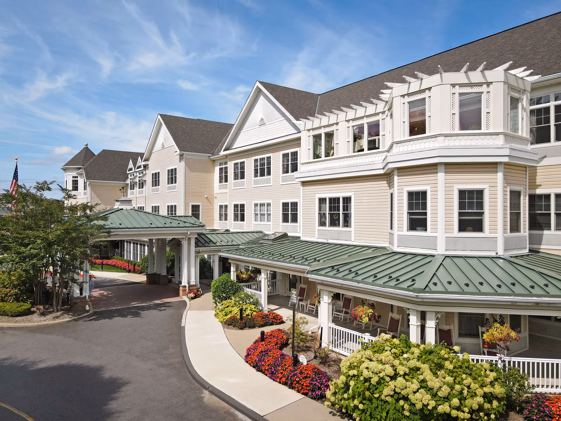 Exterior view of a large senior living facility building with beige siding and white trim, featuring a green metal roof over the entrance and porch area. The building has multiple windows and a decorative bay window section. The entrance is surrounded by well-maintained landscaping with colorful flowers and bushes under a clear blue sky.