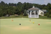 A well-maintained croquet lawn with balls and wickets set up, surrounded by green grass and trees. In the background, there is a white gazebo and a small building, with benches placed near the edge of the lawn.