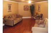 A cozy living room with two beige sofas facing each other, a wooden floor, two framed pictures on the walls, and a tall green plant in the corner. There are also two decorative chairs with cushions near the plant.