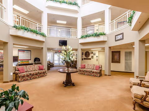 Spacious and well-lit common area in a senior living facility with beige carpet, floral-patterned sofas, a round wooden table with a large floral arrangement in the center, and multiple seating areas. The room features high ceilings with an open second-floor balcony lined with green plants and railings. There are framed pictures on the walls and a flat-screen TV mounted on the far wall.