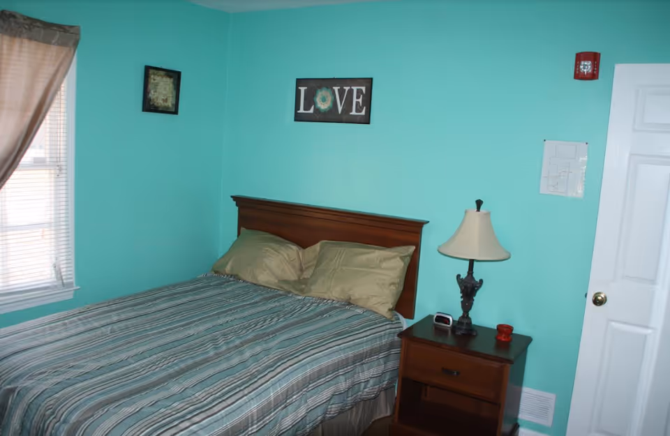 A bedroom with turquoise walls featuring a wooden bed with striped bedding and two pillows. There is a wooden nightstand with a lamp, a small clock, and a red candle. On the wall above the bed is a framed sign that says 'LOVE'. A window with blinds and a curtain is on the left side, and a white door is on the right side of the room.
