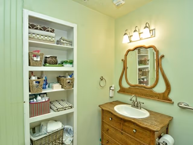 A bathroom with a wooden vanity featuring a white sink and a decorative wooden-framed mirror above it. There is a three-light fixture mounted above the mirror. To the left, there is a built-in shelving unit with various baskets, towels, and toiletries. The walls are painted light green.