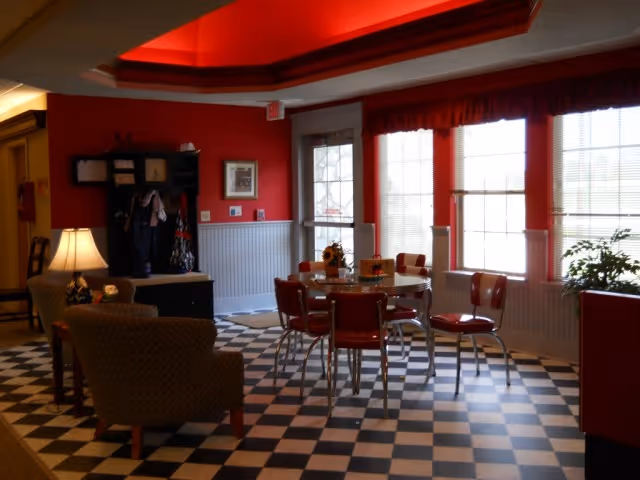 Bright red common room with a black-and-white checkered floor, a round dining table surrounded by red chairs and nearby lounge seating by windows.