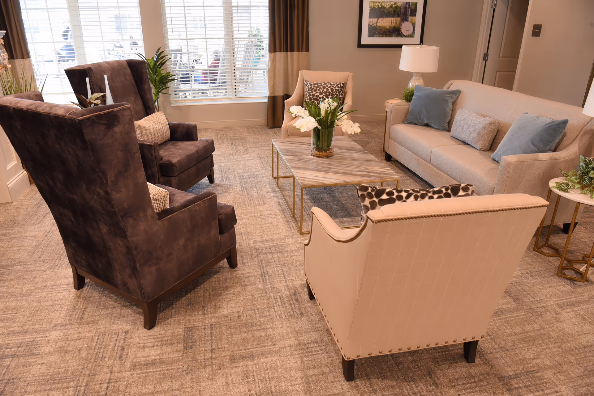 A cozy living room area with a beige sofa adorned with blue and gray pillows, two dark brown armchairs, and a light beige armchair with a patterned pillow. A rectangular coffee table with a vase of white flowers sits in the center on a patterned carpet. The room has large windows with blinds and curtains, a side table with a lamp and plant, and a framed picture on the wall.