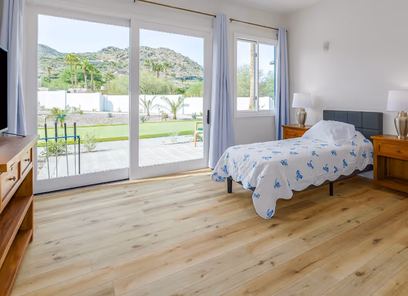 Bright single-occupancy bedroom with a bed, wooden nightstands and sliding glass doors opening to a garden and mountain view.