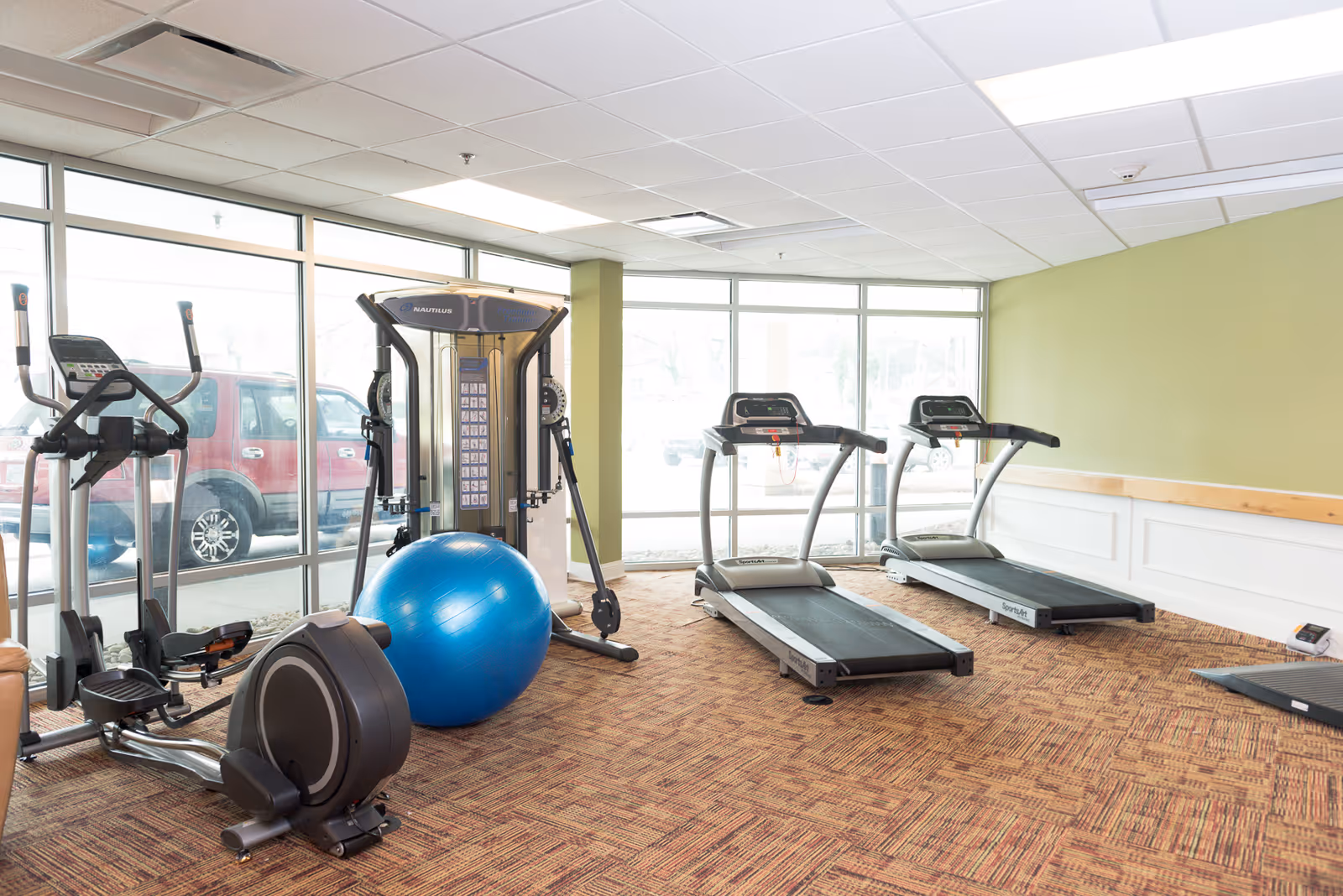 A fitness room with exercise equipment including two treadmills, an elliptical machine, a rowing machine, a multi-function weight machine, and a large blue exercise ball. The room has large windows letting in natural light and a green accent wall.