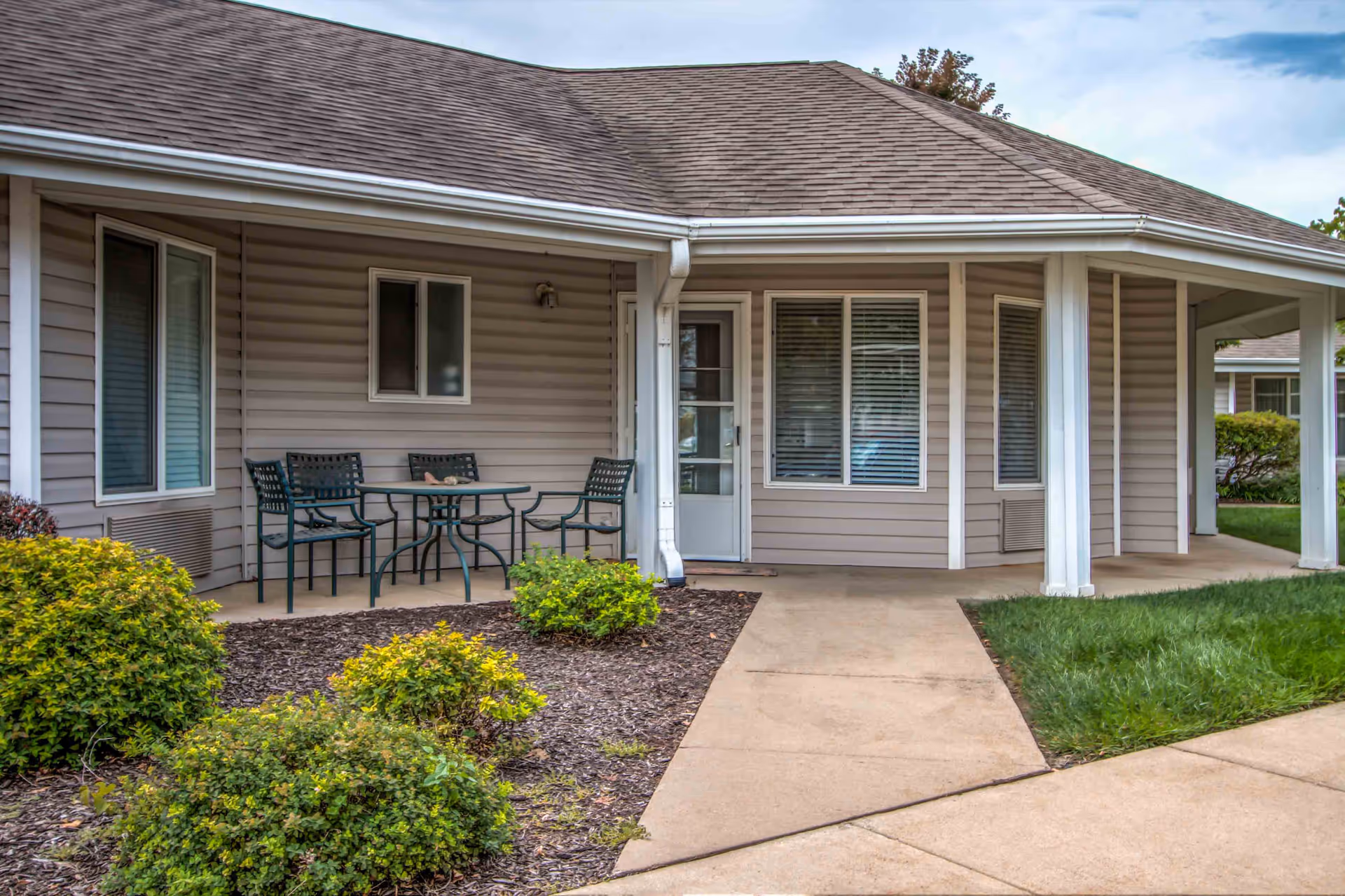 Outdoor patio area of a residential building with beige siding, a small table with four chairs, and surrounding green bushes and grass under a partly cloudy sky.