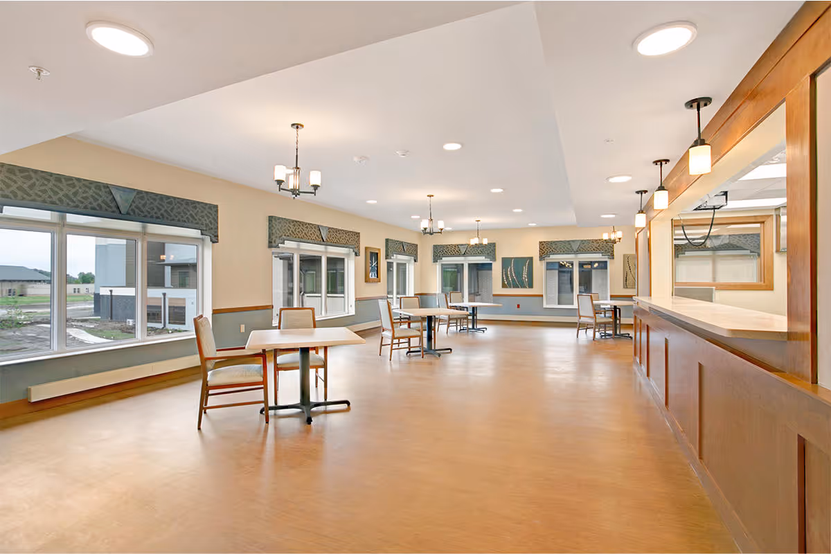 Bright open dining/activity room with several tables and chairs, large windows on the left, and a wooden serving counter on the right.