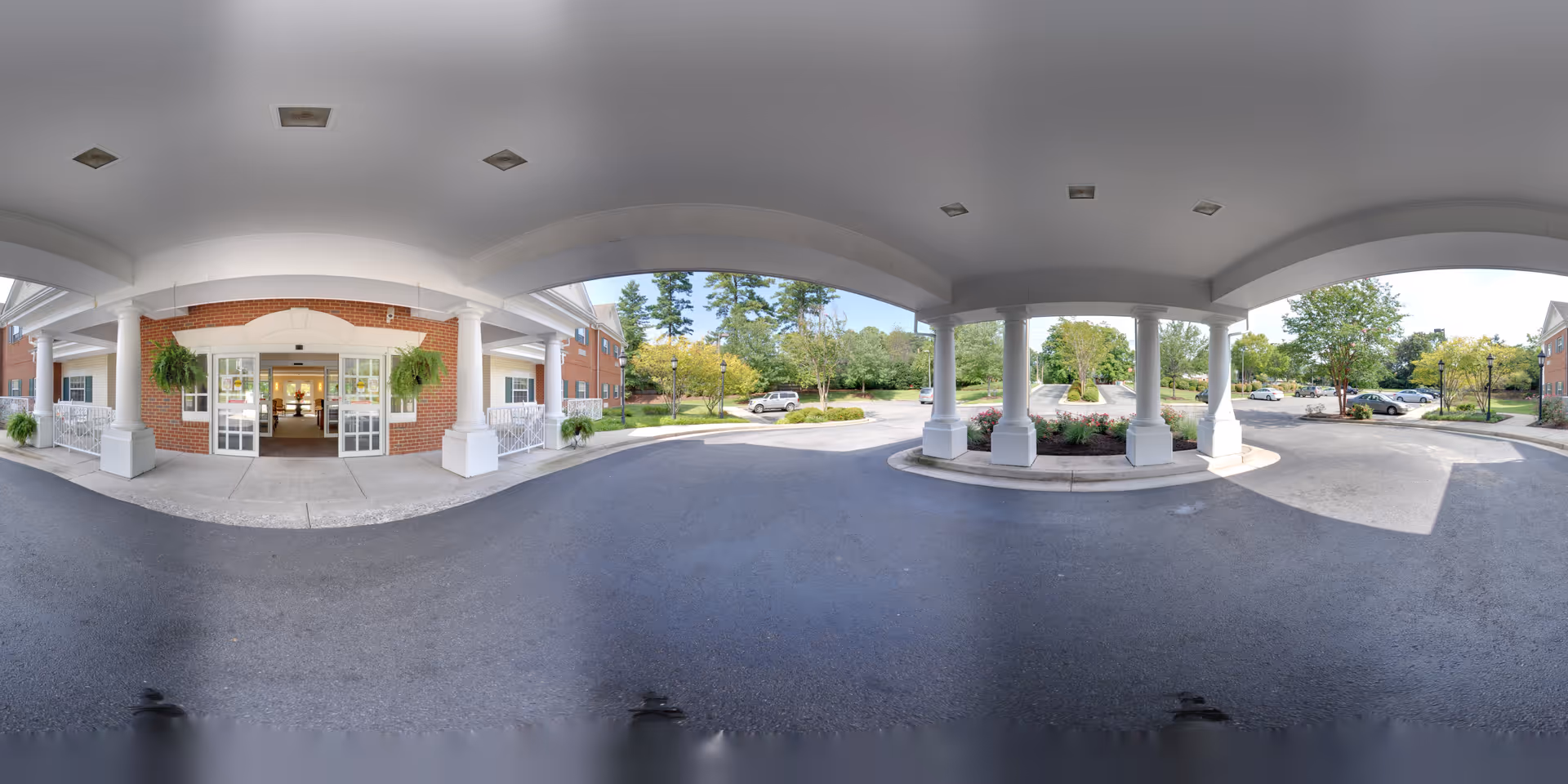 Covered porte-cochere entrance to a brick senior living building with white columns, hanging plants, and a circular drive.