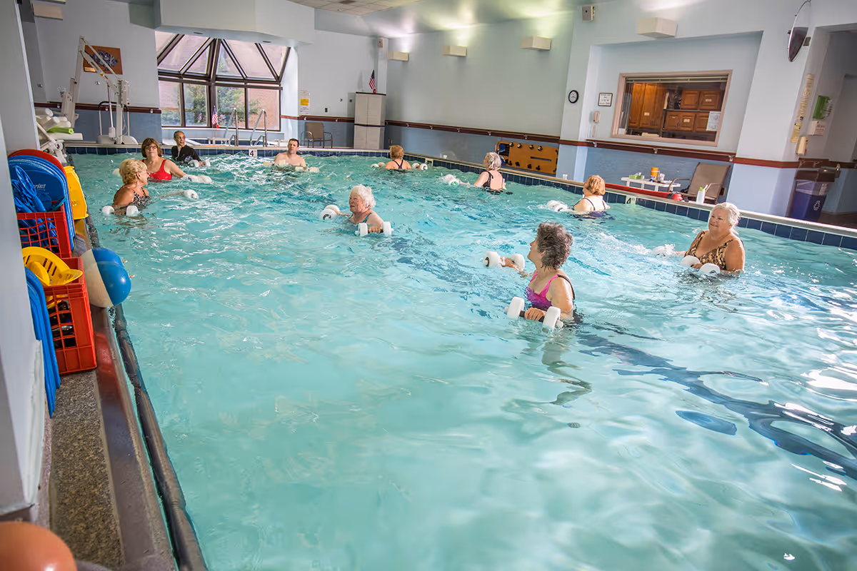 A group of elderly people participating in a water exercise class in an indoor swimming pool at a senior living community. The pool area has large windows, light blue walls, and various pool equipment along the side.