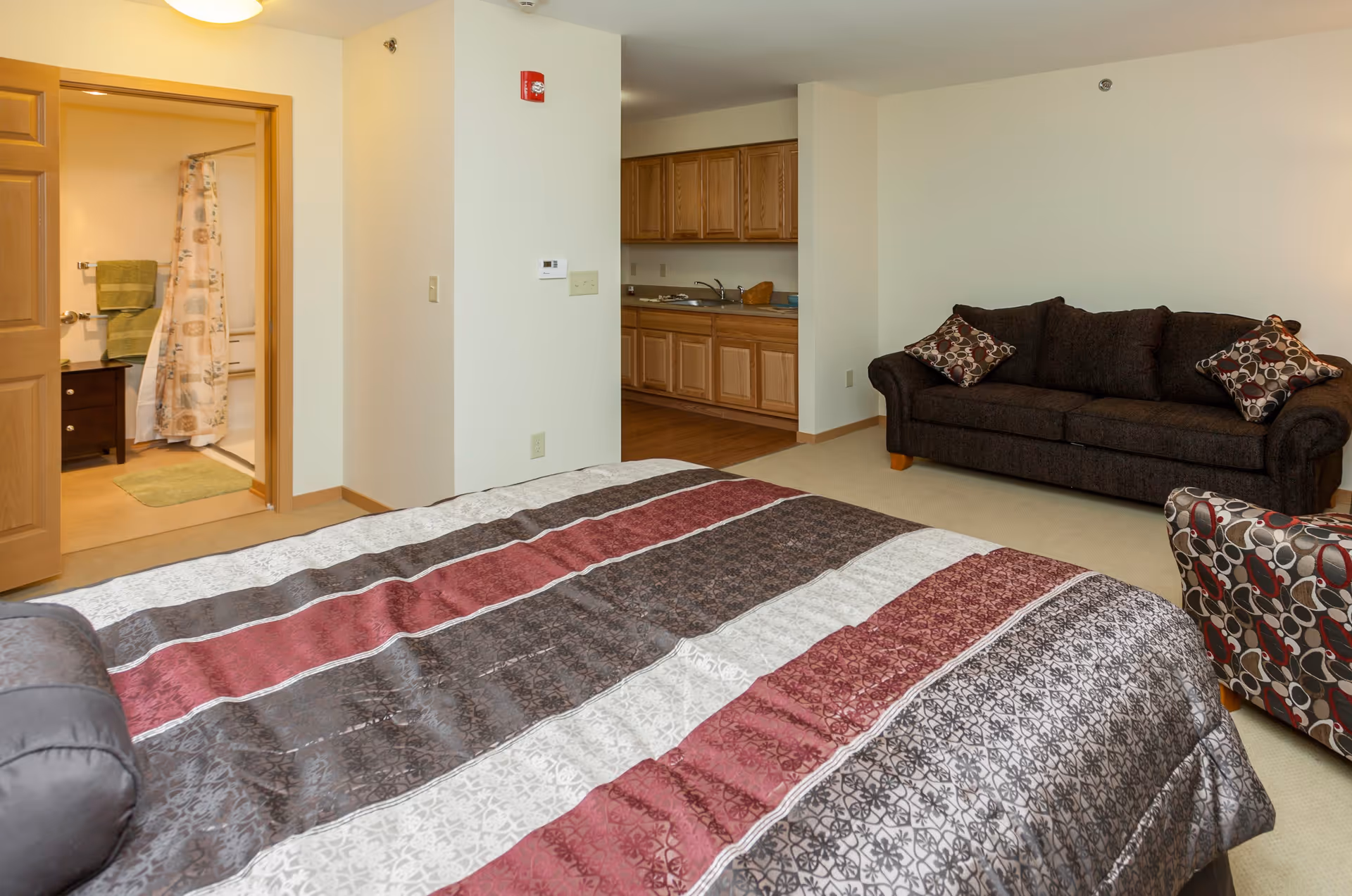 Interior view of a senior living facility room at Edencrest at Riverwoods showing a bed with a patterned bedspread in the foreground, a dark brown couch with patterned pillows, a matching armchair, a kitchenette with wooden cabinets, and a bathroom with a shower curtain and green towels visible through an open door.