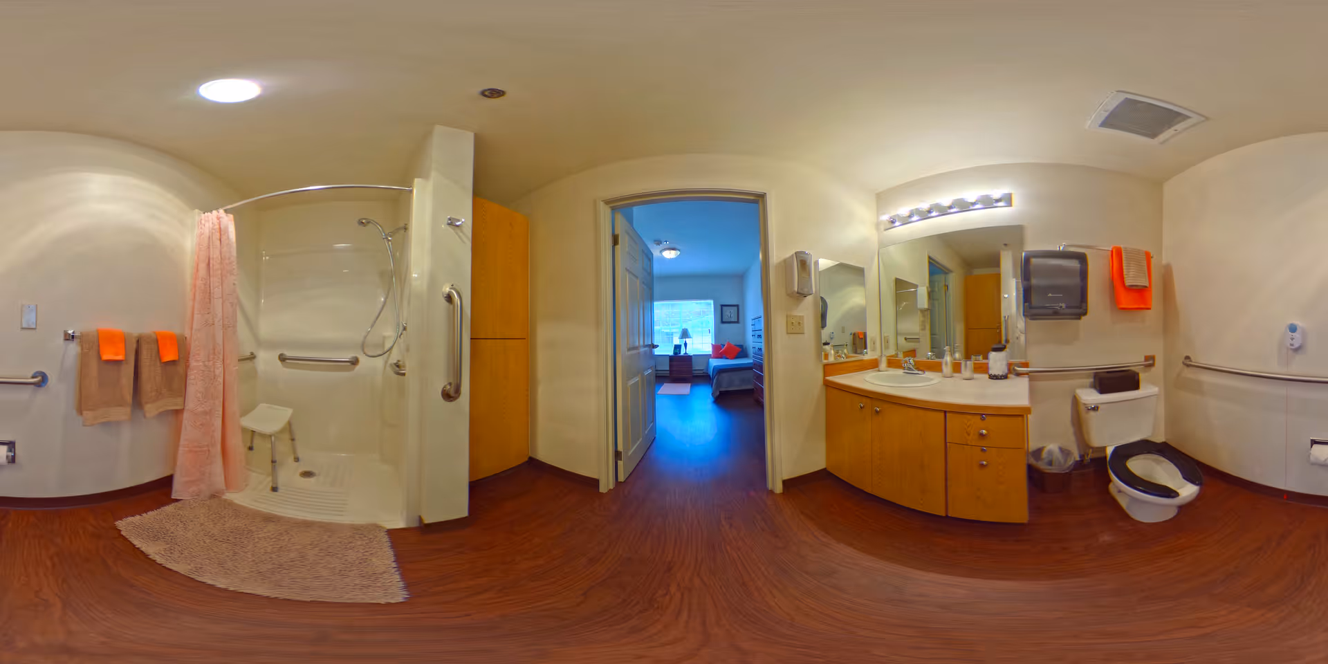 A spacious senior living bathroom with a walk-in shower equipped with a shower chair and grab bars, a toilet with grab bars, a sink with a large mirror, and orange and beige towels hanging on the walls. The bathroom has wood-style flooring and light-colored walls. Through an open door, a bedroom with a bed and window is visible.