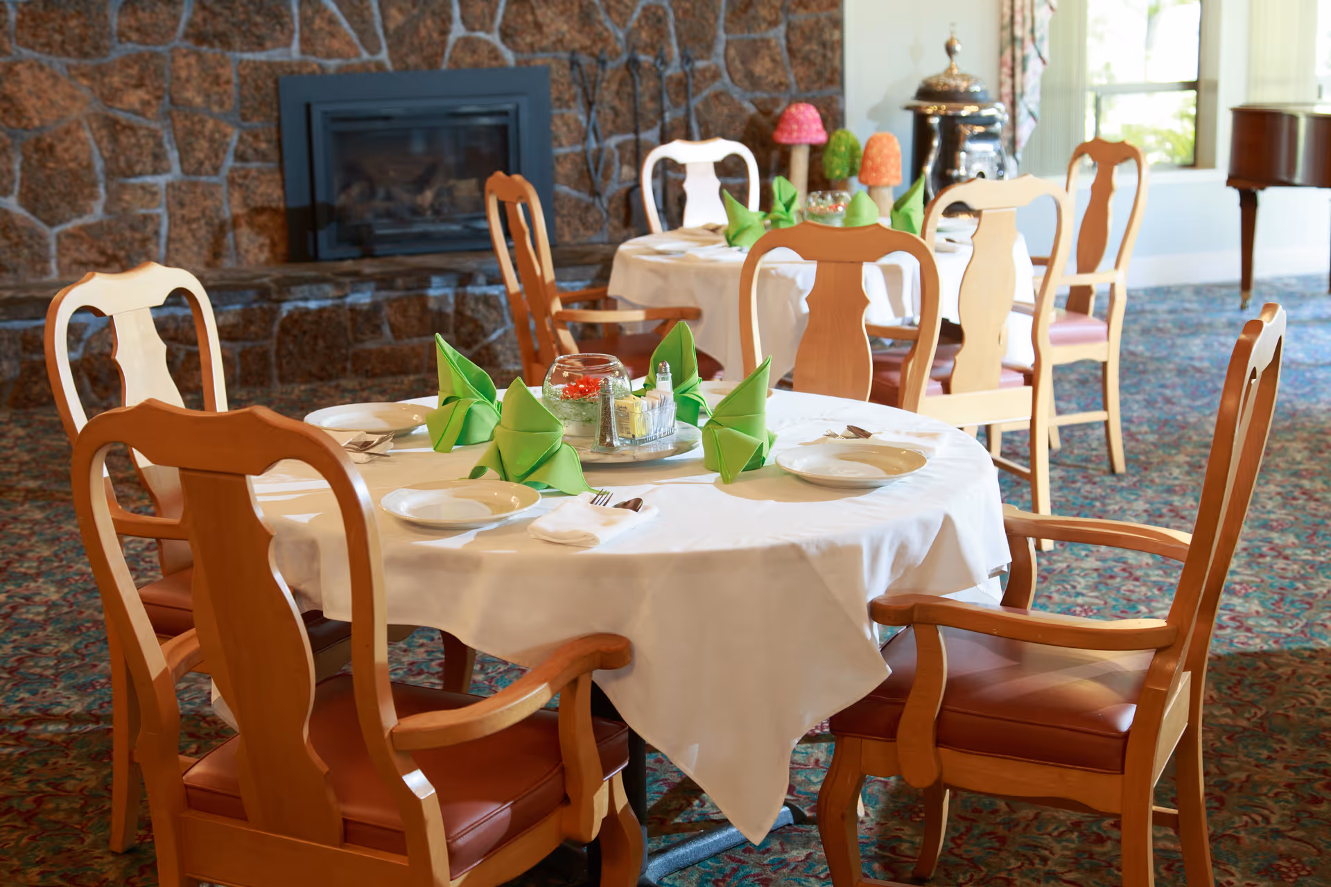 A dining room with round tables covered in white tablecloths, set with plates, utensils, and green folded napkins. Wooden chairs with armrests surround the tables. In the background, there is a stone fireplace and large windows letting in natural light.