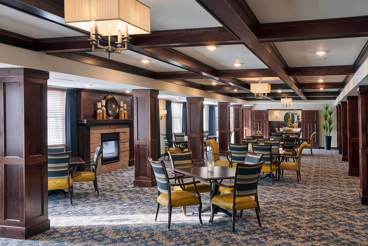 A spacious dining room with multiple tables and chairs featuring yellow cushions and striped backs. The room has wooden columns, a patterned carpet, a fireplace with decorative candles, large windows with blinds and curtains, and ceiling lights with rectangular lampshades.