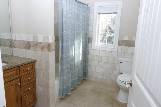 Bathroom with beige tiled walls and floor, a wooden vanity with granite countertop and sink on the left, a white toilet on the right, a window above the toilet, and a shower with a blue patterned curtain in the center.