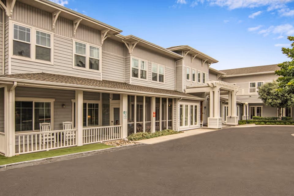 Exterior view of a two-story senior living facility building with beige siding, multiple windows, a covered porch with rocking chairs, and a paved driveway under a clear blue sky.