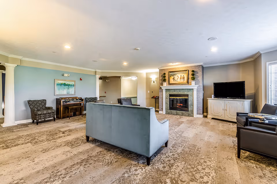 A spacious living room in an assisted living facility featuring a light blue sofa, two patterned armchairs, a fireplace with a painting above it, a TV on a white cabinet, and a piano against a light blue wall. The room has beige patterned carpet and soft lighting.