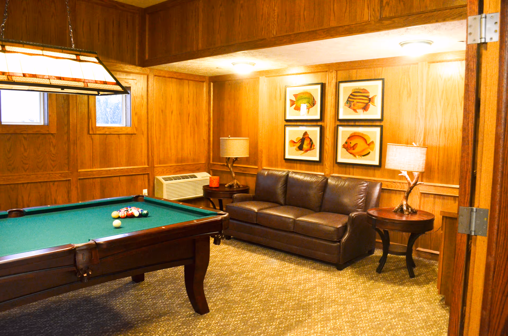 A cozy game room with wood-paneled walls featuring four framed fish illustrations. The room includes a green felt pool table with billiard balls set up, a brown leather sofa, two round wooden side tables each with a lamp, and two small windows near the ceiling.
