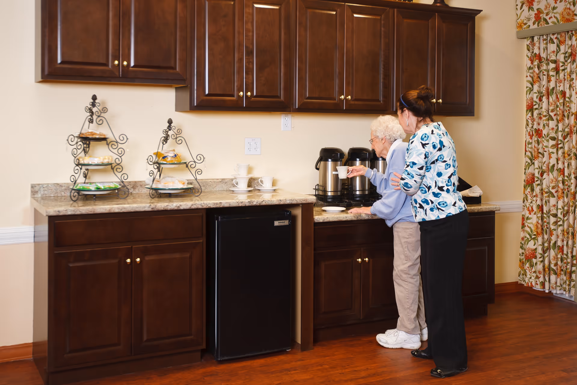 An elderly woman and a caregiver stand at a kitchen counter with dark wooden cabinets and a granite countertop. The elderly woman is holding a cup near coffee dispensers, while the caregiver supports her. There are tiered trays with snacks and stacked cups on the counter. Floral curtains are visible on the right side.