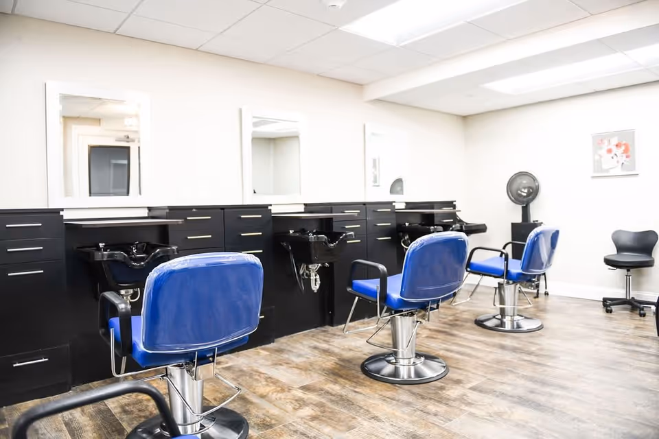 Interior of a salon area with three blue salon chairs in front of black counters with sinks and mirrors. The room has wood flooring, white walls, and a ceiling with fluorescent lighting. There is also a black rolling stool and a hair dryer in the corner.