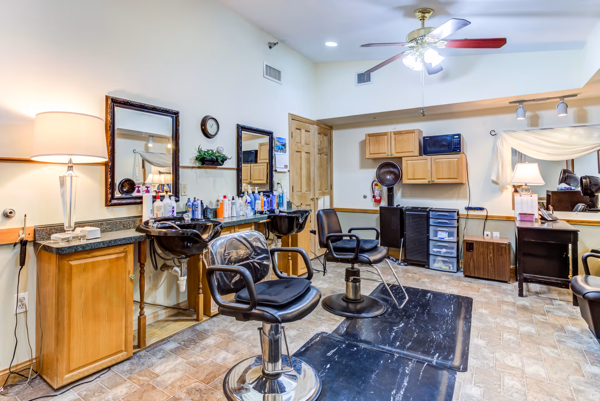 Interior view of a hair salon area in an assisted living facility with two black salon chairs, two black hair washing sinks, mirrors on the wall, various hair care products on the counter, wooden cabinets, a ceiling fan with lights, and a lamp on a countertop.