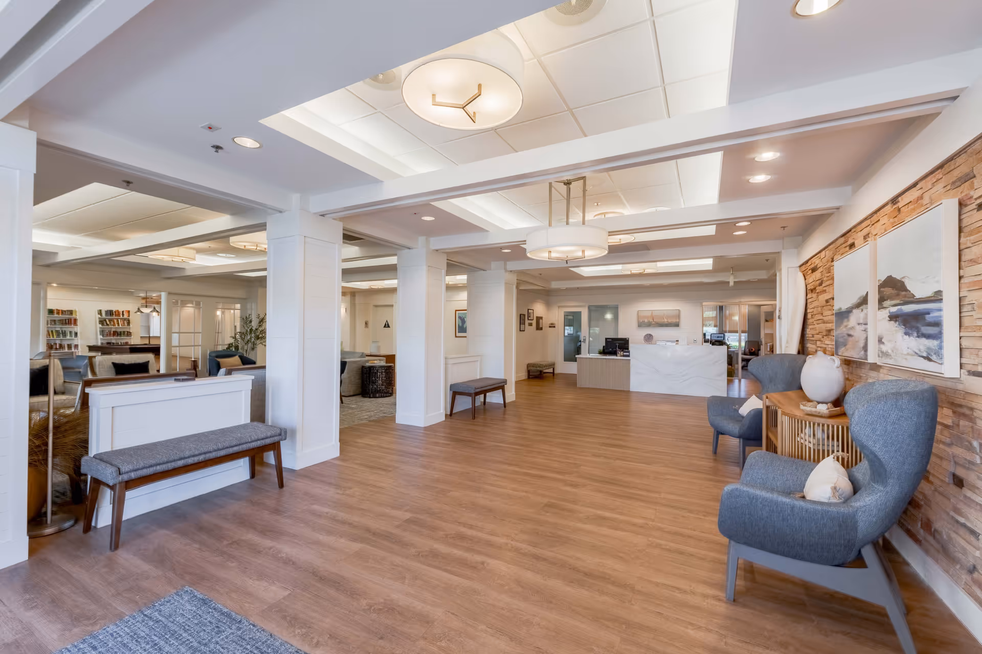 Spacious and well-lit senior living facility lobby with wooden flooring, white pillars, modern ceiling lights, a reception desk in the background, and comfortable seating including blue armchairs and benches. The right wall features a stone accent with framed artwork and decorative items on a small table.
