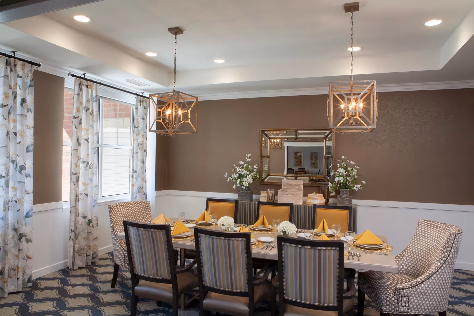 A well-lit dining room with a rectangular table set for eight people. The table has yellow napkins, plates, glasses, and floral centerpieces. There are patterned curtains on the windows, two modern pendant lights hanging from the ceiling, and a large mirror on the brown accent wall reflecting the room.