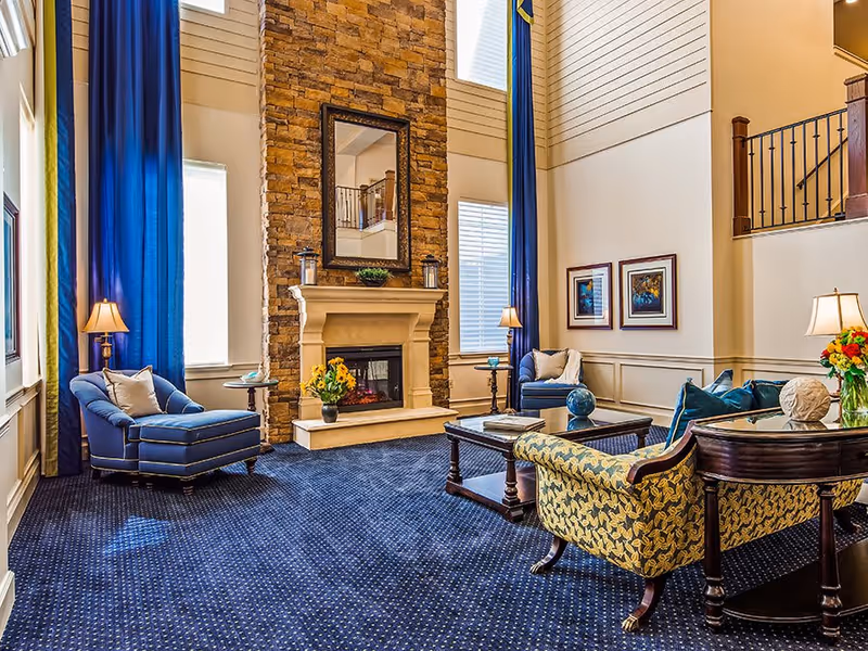 A spacious and elegant living room with high ceilings, featuring a stone fireplace with a large mirror above it, blue carpet, blue curtains, and comfortable seating including a blue armchair, a patterned yellow and blue sofa, and a wooden coffee table with decorative items. There are also two framed pictures on the wall and several lamps providing warm lighting.