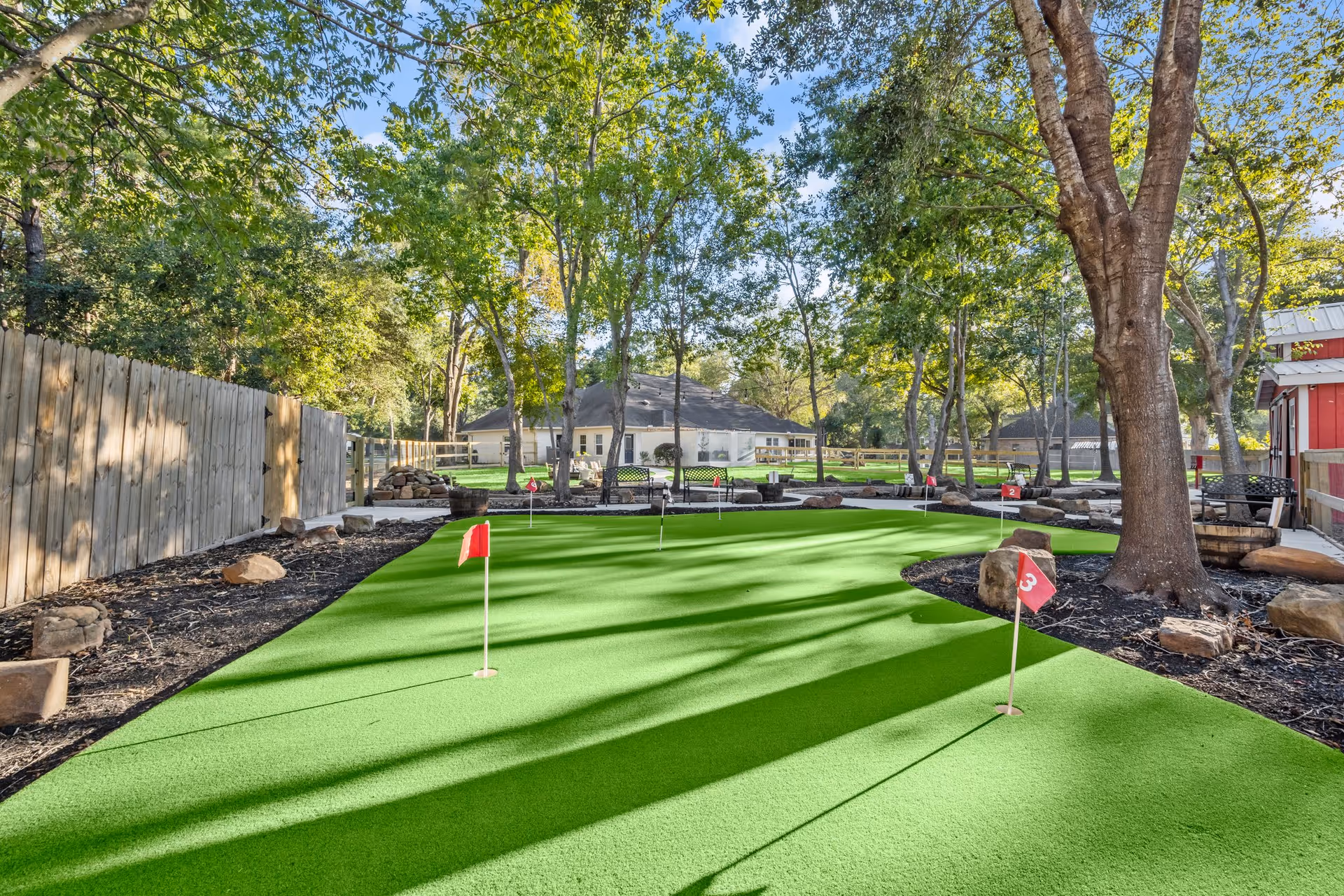 Outdoor putting green with several small red flags numbered 2 and 3, surrounded by trees, rocks, benches, and a wooden fence, with a building visible in the background under a clear blue sky.