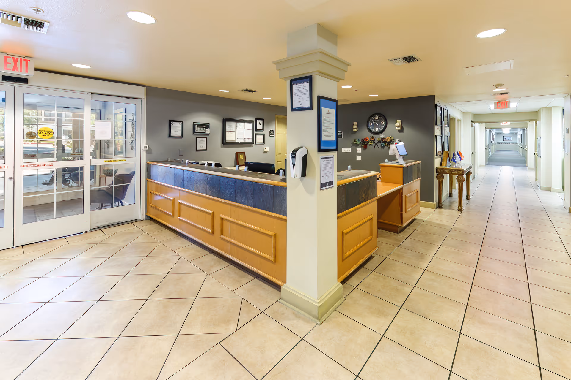 Interior view of a senior living facility reception area with a wooden front desk, tiled floor, and a long hallway extending to the right. The reception desk has a dark countertop and is positioned around a central column with notices posted on it. There are framed certificates and a clock on the wall behind the desk. Glass double doors with exit signs are visible on the left side.