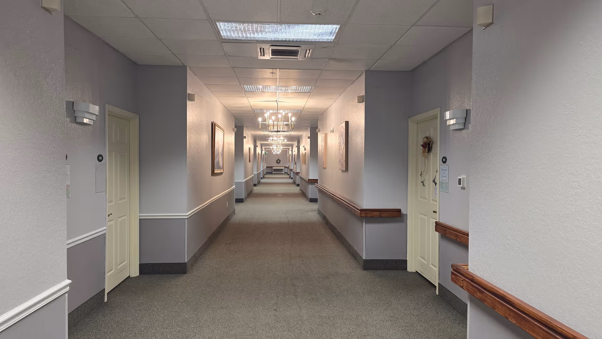 Long carpeted interior hallway in an assisted living facility with doors on both sides, handrails, chandeliers, and framed artwork.