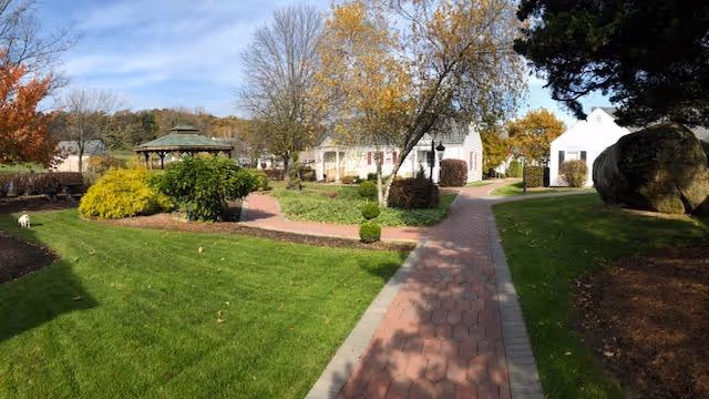 A landscaped outdoor area with a paved walkway leading through green grass and trees with autumn foliage. There is a gazebo on the left side and several buildings in the background under a partly cloudy sky.
