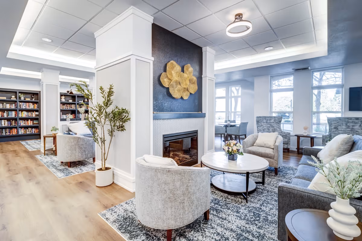 A bright and spacious living room area with modern furniture including a gray sofa, two armchairs, a round coffee table with a flower arrangement, and a fireplace with decorative golden wall art above it. The room features large windows letting in natural light, wooden flooring, a patterned rug, and bookshelves filled with books in the background.
