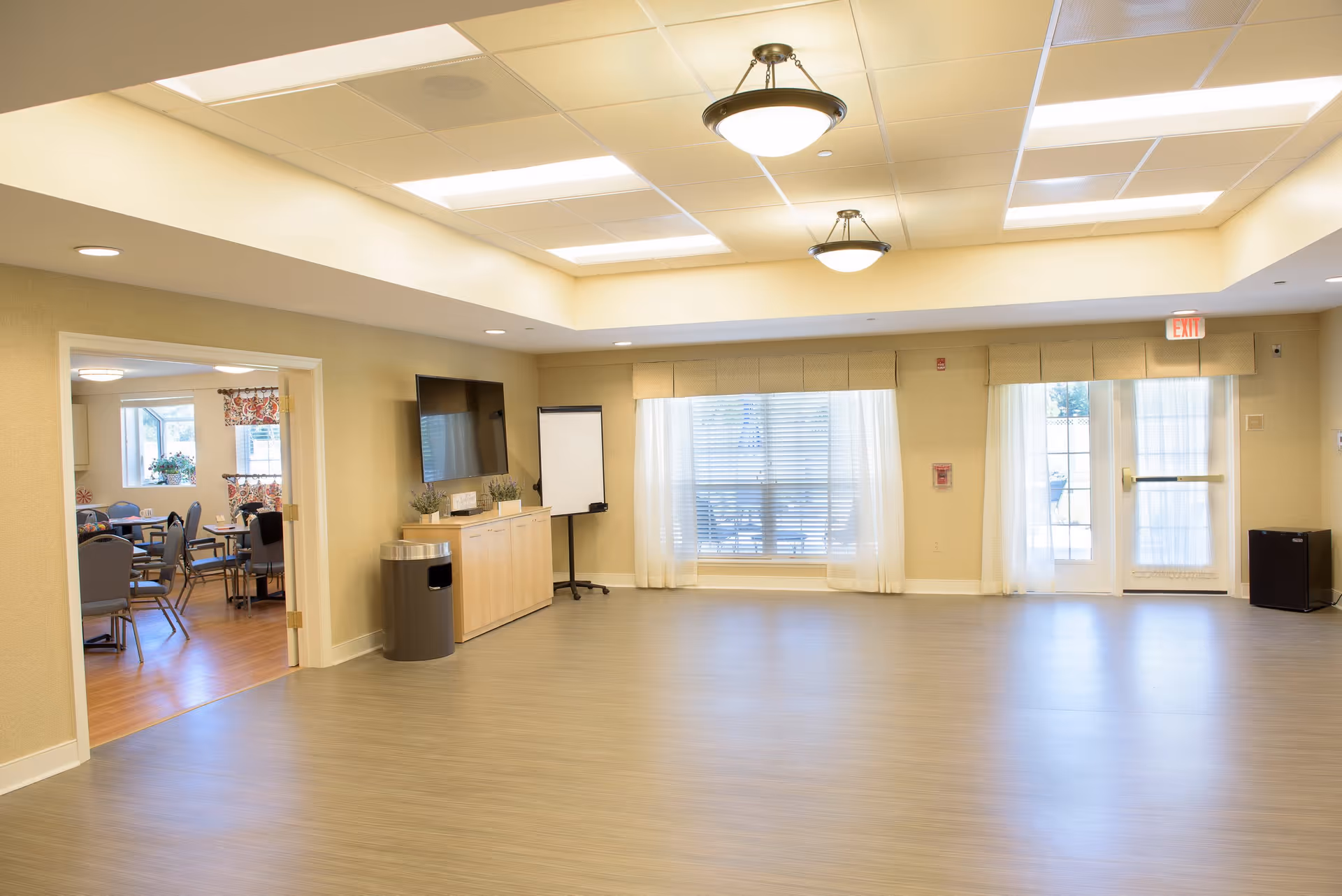 A spacious, well-lit common room with light-colored walls and flooring. The room features two ceiling light fixtures, large windows with sheer curtains, and a glass door leading outside. There is a wooden cabinet with a flat-screen TV mounted above it, a trash can, and a flip chart stand. To the left, an open doorway leads to a dining area with tables and chairs.