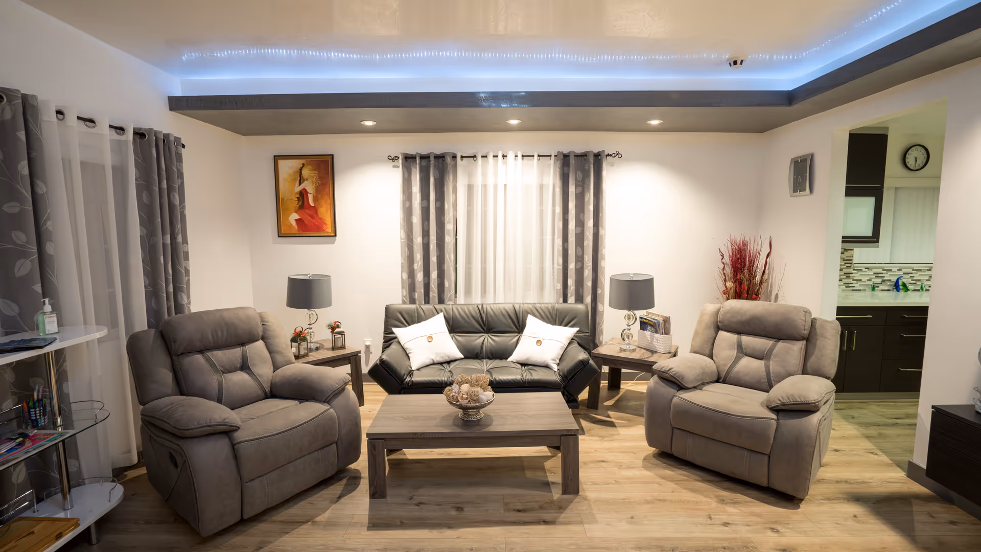 Well-lit living room with a central dark sofa, two gray recliners, a wooden coffee table and side tables under recessed ceiling lighting.