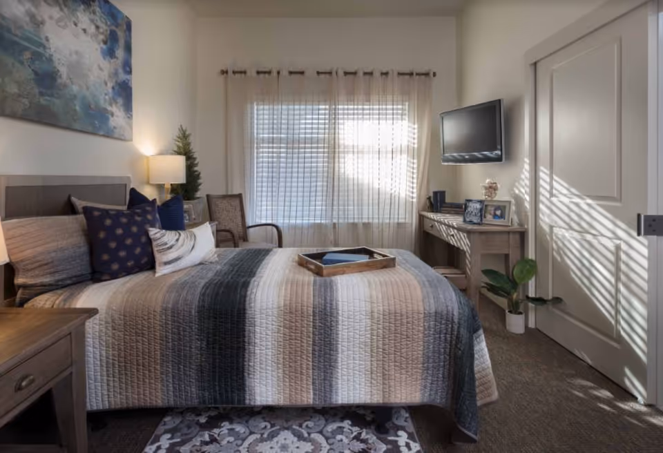 A cozy bedroom with a neatly made bed featuring a striped quilt and multiple pillows. There is a wooden nightstand with a lamp on the left side of the bed. A window with sheer curtains allows natural light to fill the room. A wooden desk with framed photos and decorative items is positioned against the right wall beneath a mounted flat-screen TV. A chair and a small potted plant are also visible in the room.