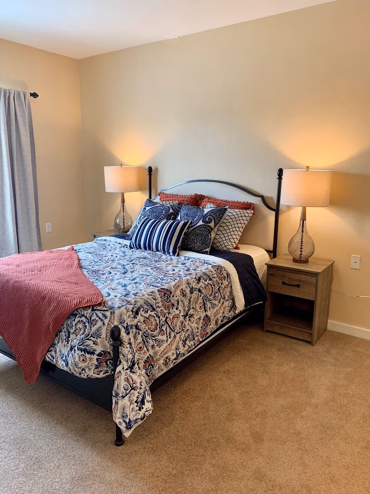 A neatly made bed with a black metal frame in a bedroom. The bed has a floral patterned comforter, multiple decorative pillows in various patterns and colors, and a red throw blanket draped over the foot of the bed. On each side of the bed are wooden nightstands with glass-based lamps that are turned on. The room has beige walls, beige carpet, and a window with gray curtains on the left side.