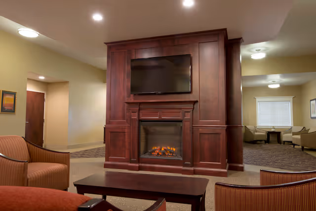 A cozy communal lounge featuring a large wooden fireplace unit with an electric fire and a wall-mounted TV, surrounded by upholstered chairs and tables.