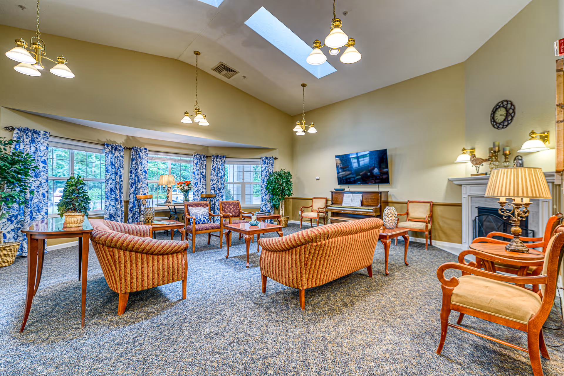 A spacious and well-lit living room with multiple seating options including striped sofas, wooden chairs with cushions, and small tables. The room features large windows with blue and white patterned curtains, a piano with sheet music, a wall-mounted TV, a fireplace, and several lamps and plants. The ceiling has skylights and hanging light fixtures.