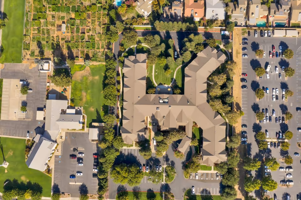 Aerial view of Dale Commons senior living facility showing a large building with a complex roof structure surrounded by trees, parking lots filled with cars, and adjacent green spaces including a garden area.