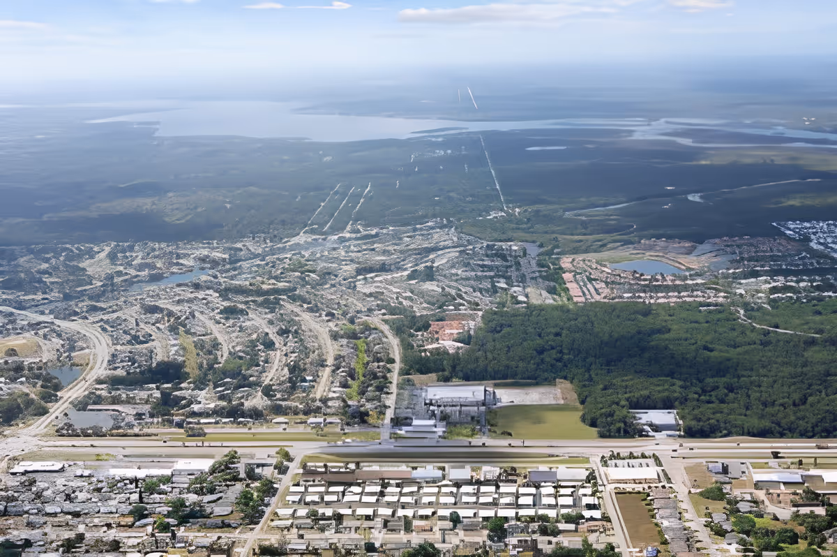 Aerial view of a coastal landscape showing residential neighborhoods, roads, wooded areas, and a large body of water on the horizon.
