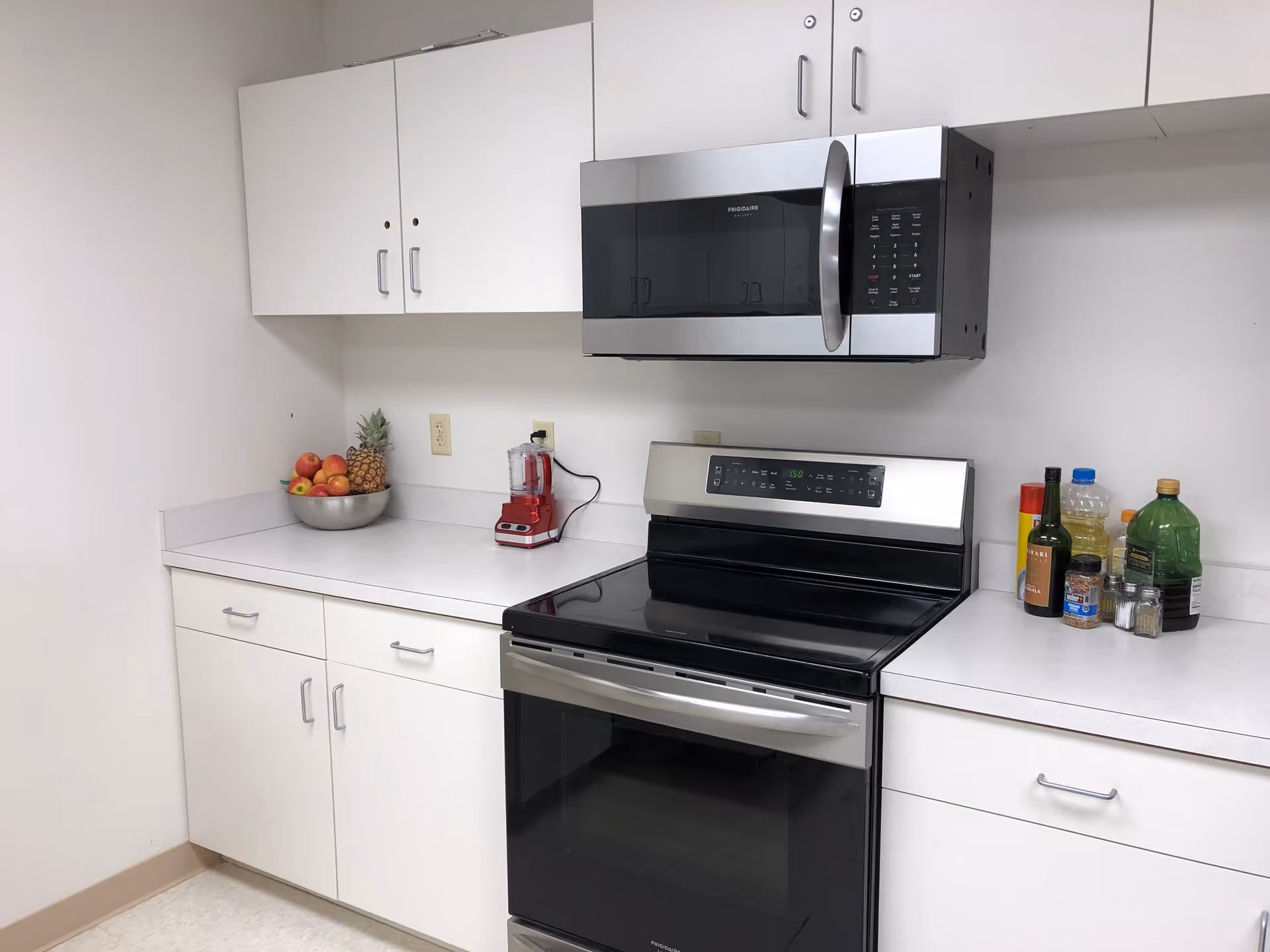 A clean kitchen area with white cabinets and countertops. There is a stainless steel microwave mounted above a black and stainless steel electric stove. On the left countertop, there is a bowl of fruit including apples and a pineapple, and a red blender. On the right countertop, there are various bottles and containers including cooking oil, vinegar, and spices.