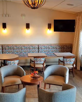 Small communal dining and lounge area with round tables, upholstered chairs, a patterned banquette, wall sconces, and a mounted TV.
