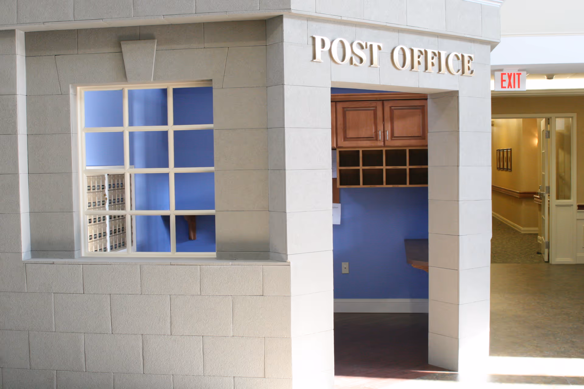 Interior view of a post office area within a facility, showing a stone-like wall with a window opening revealing mailboxes inside. Above the entrance is a sign that reads 'POST OFFICE'. To the right, a hallway with an exit sign and open doors is visible.
