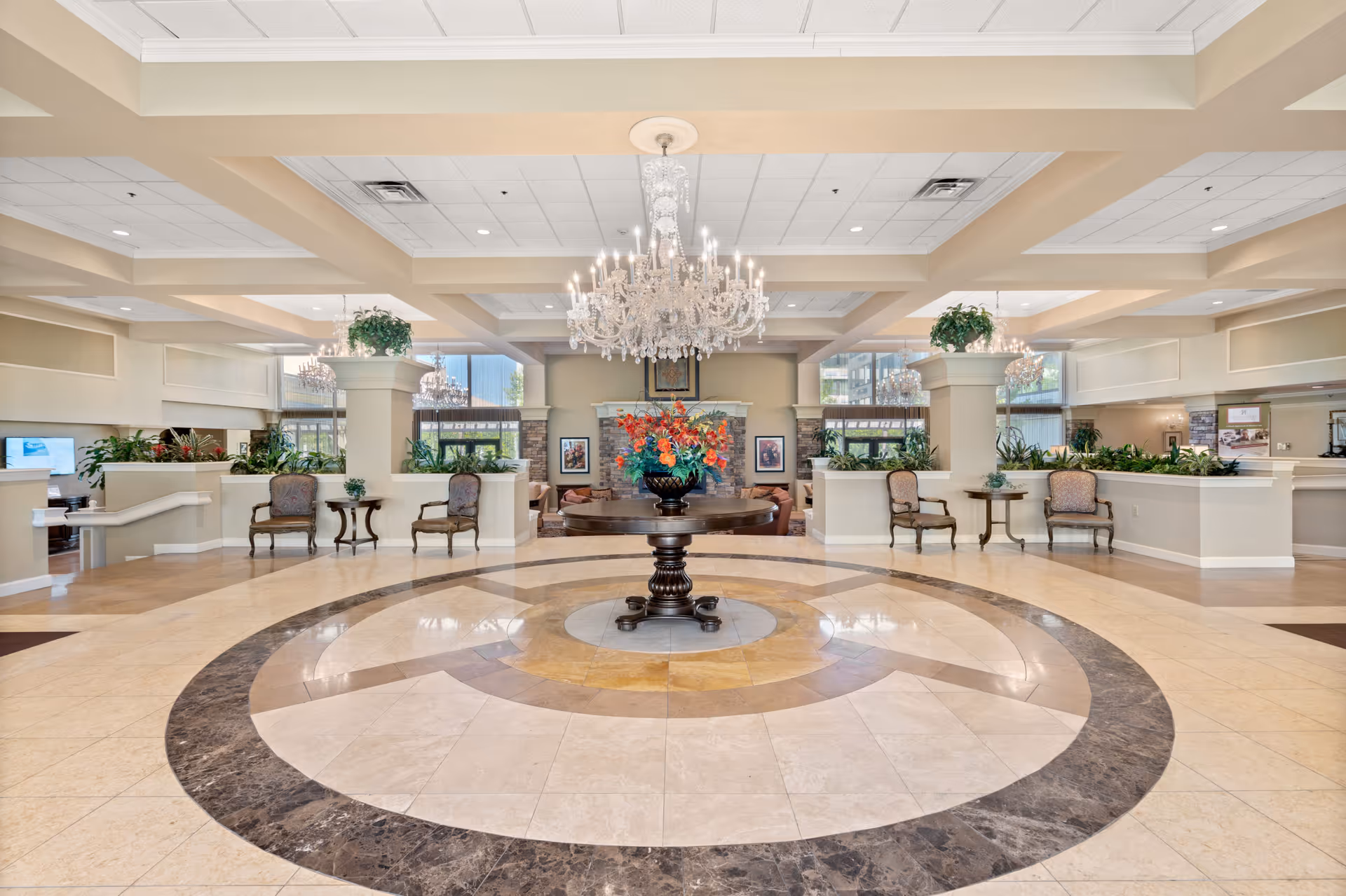 Spacious ornate lobby with a circular marble floor medallion, central table holding a floral arrangement, chandeliers, seating areas and plants.